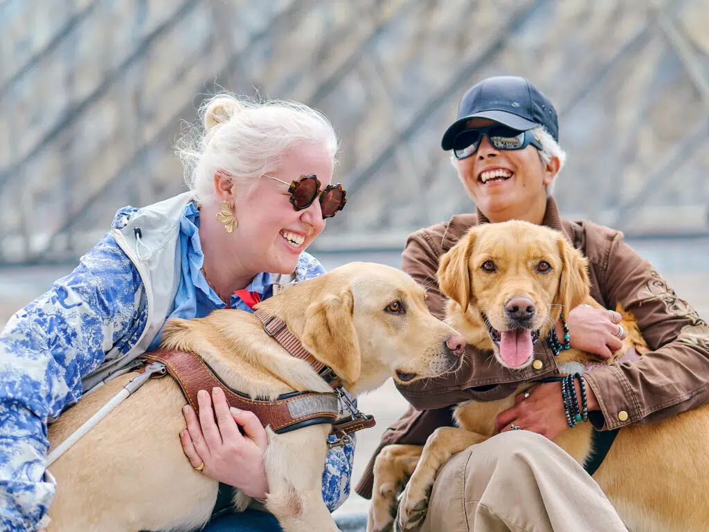 Photo de deux binômes avec leur chien guide. La photo est prise devant le Louvre.