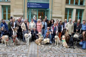 Photo de groupe de tous les chiens guides retraités en 2025, accompagnés de leurs anciens maîtres. La photo est prise en extérieur.
