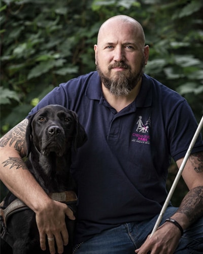 William est assis à l'École de Chiens Guides de Paris. Il est avec un labrador noir qui porte son harnais et tient une canne d'éducation.