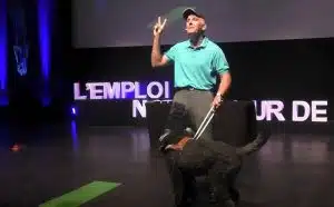 Norbert est sur scène avec son chien guide Leroy, il fait un signe de paix avec ses doigts.
