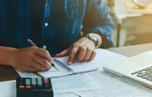 Photo d'un homme qui tient dans sa main un stylo et devant lui ce trouve un cahier et une calculette