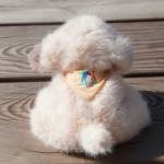 Photo d’une peluche couleur sable, de style Fluppy, vue de dos. Elle porte un bandana jaune sur lequel est brodé le logo de l’École.