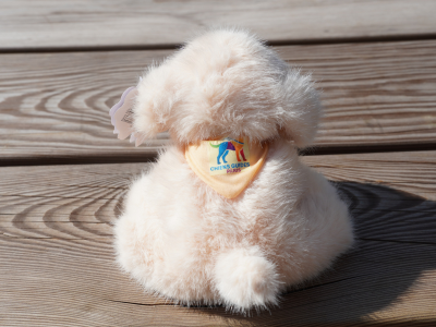 Photo d’une peluche couleur sable, de style Fluppy, vue de dos. Elle porte un bandana jaune sur lequel est brodé le logo de l’École.
